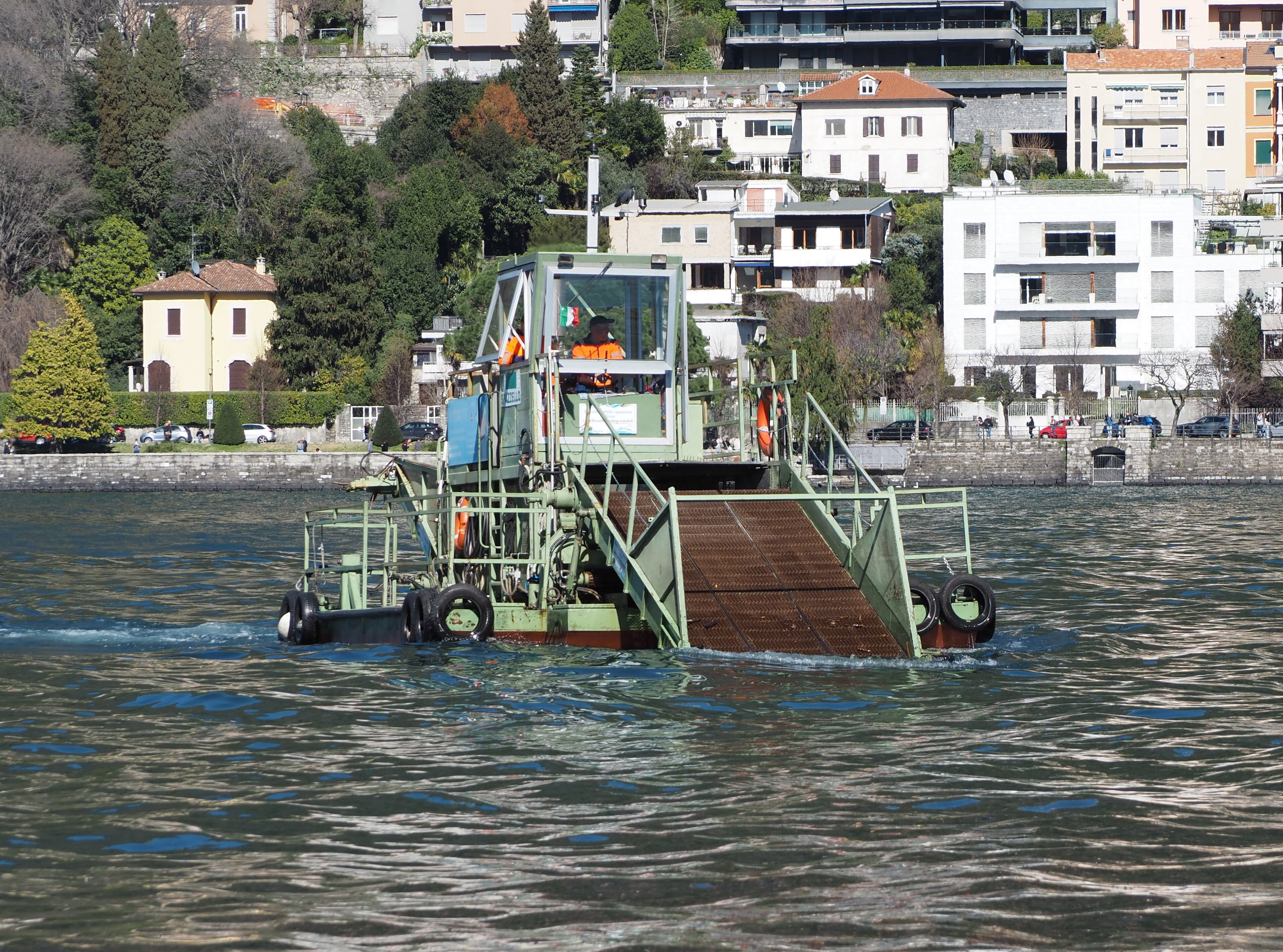 Pistonheads - The image shows a green barge on the water. The barge is equipped with a crane, suggesting it's used for heavy lifting or transportation. There are two individuals visible on the boat, likely operators or crew members. In the background, there are residential buildings and a body of water, indicating that this scene might be taking place near a shoreline or in a marina.