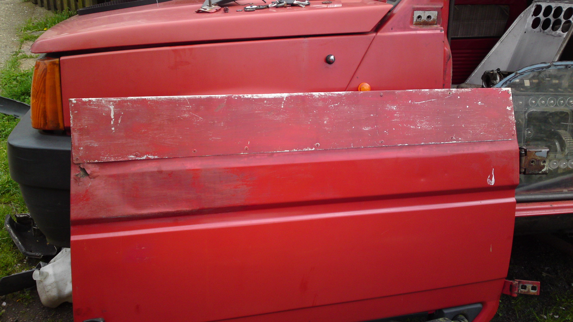 An old suitcase is sitting on the ground - Pistonheads - This image features a close-up view of a red vehicle appearing to be a truck. The truck is parked so we see its side profile. A distinctive feature of the truck is the absence of the driver's side door, indicating it is either broken or missing this part of its vehicle structure. Additionally, on the driver's side door, there is a large square of metal or wood with a rough, weathered texture, which has been removed from the vehicle. The backdrop suggests the photo is taken outdoors, with bits of foliage and possibly a fence.