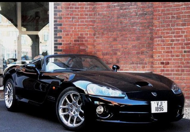 Hi Guys im looking for another Viper  - Page 5 - Vipers - PistonHeads - This image captures a sleek, black sports car with a convertible top, parked on the side of a road. The car is positioned in front of a large, red brick wall, suggesting a contrast between the vehicle's streamlined design and the wall's sturdy, traditional architectural style. The photograph conveys a sense of drama, enhanced by a shallow depth of field that highlights the car as the focal point. The perspective is from an angle that accentuates the car's profile and gives a glimpse into the interior through the open door and light reflected on the car's bodywork.