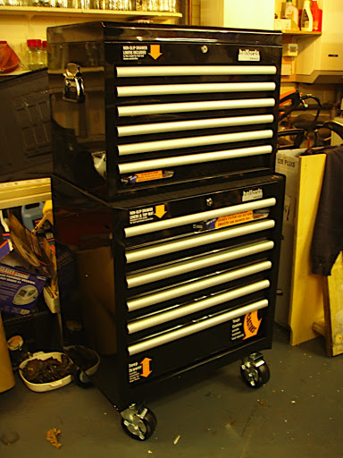 A pile of luggage sitting on top of a wooden floor - Pistonheads - The image shows a black and white tool chest with sturdy wheels, sitting on a garage-like floor. The chest has multiple drawers, with at least four highlighted by triangular stickers on the top drawer. It is equipped with locks and bearings on the bottom, suggesting mobility and security features. To the left of the chest, there is clutter, including household items and papers, indicating that the area is likely utilitarian rather than pristine. An extension cord is visible, further emphasizing the industrial feel of the space.