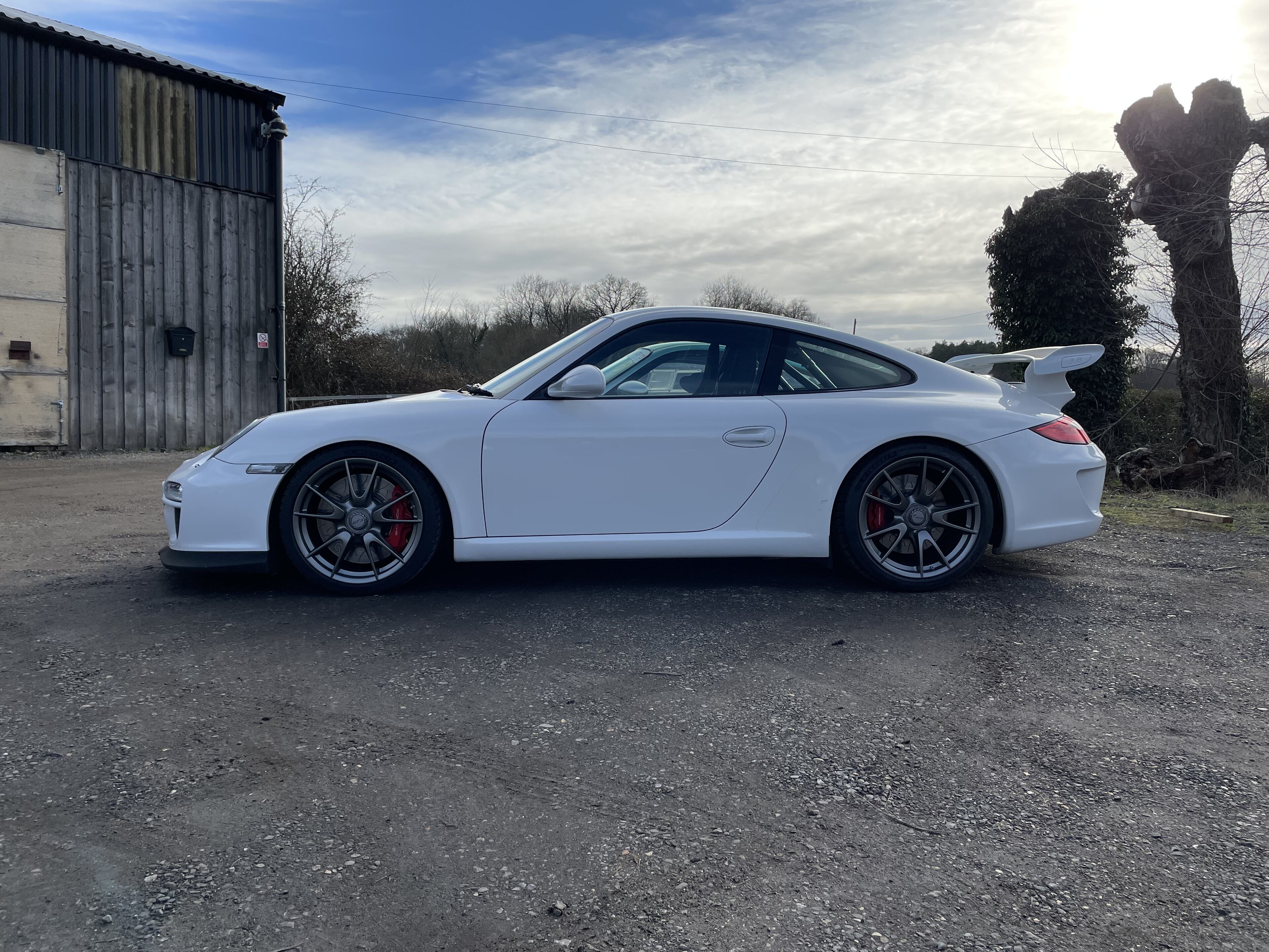 Pistonheads - The image showcases a white Porsche parked on the side of a road. The car is positioned next to a building, under a clear sky. It appears to be a sunny day, with sunlight illuminating the vehicle and creating reflections on its surface. The car's design features sleek lines and a distinctive front grille, indicative of Porsche's sporty aesthetic. The Porsche logo is prominently visible on the hood. In the background, there's an old tree, adding a touch of nature to the urban setting. There are no other objects or people in the immediate vicinity of the car, giving it center stage in this composition.