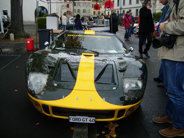 Meeting October Pistonheads Morges British - The image depicts a scene at a sports car show. A black and yellow race car is parked on a blacktop street. A person is walking by, holding a camera, likely capturing the event. In the background, several other people can be seen, some of them also walking and others standing. The car is adorned with a sign stating "www.cameroncompany.com", and there's a ticket on the windshield, indicating that it's part of the event.