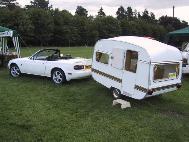 A white and blue truck parked in a field - Pistonheads - The image depicts a camping scene with two main elements. On the left is a white Japanese sports car, specifically a Nissan GT-R, that has been converted into a race car track. On the right is a vintage-style Airstream camper, painted white with the Stellamann logo, standing on a trailer behind the car. Both vehicles are parked on grass, which is bordering a wooded area. The overall scene suggests that the car and camper are ready for an outdoor adventure or event.