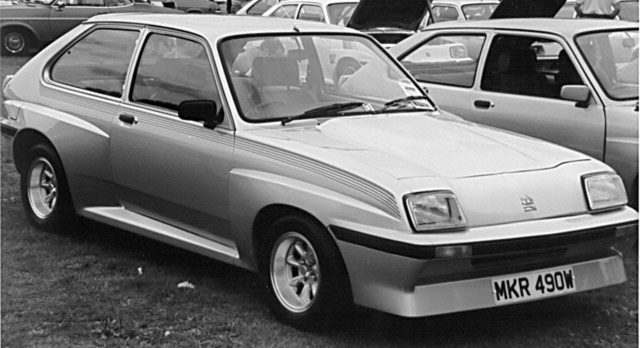 Dissapearence Pistonheads Sad Classic - The image shows a classic, compact silver car parked among other cars, possibly at a car show or exhibition. The car features a prominent black stripe running along its side, and has a unique headlight design. The vehicle's licence plate is clearly visible, displaying a combination of upper and lower case letters and numbers, indicating it may be a vintage model. The overall atmosphere of the image suggests a gathering of car enthusiasts, emphasizing the classic and distinctive style of the parked car.