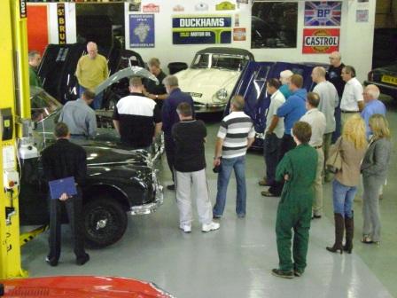 Pictures from Paul Roach's Open Day - Page 1 - Classic Cars and Yesterday's Heroes - PistonHeads - The image depicts a group of people gathered in a garage or workshop, centered on the inspection of classic cars. They seem to be examining one car in particular, which is the main focus of the scene. The setting appears to be commercial or industrial, with several other cars visible in the background. The attire of the people is casual, suggesting a relaxed, non-professional gathering. The purpose of this gathering is not explicitly clear from the image, but it might be an event, a meetup, or perhaps a repair or maintenance session.