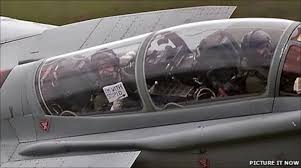 Mach Loop Typhoon ‘Incident’. - Page 4 - Boats, Planes & Trains - PistonHeads - The image shows an aircraft with two distinctive openings in its side, resembling a cockpit. There are three pilots visible, each focused inside their respective compartments. One of the pilots is holding up a sign with visible text, although the text is partially obscured. The aircraft appears to be of a modern design with sleek lines and a glossy exterior, likely a fighter jet given the visible communications equipment. There is a watermark in the bottom right corner of the image that reads "pictureitnow".