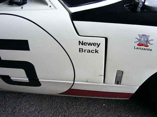 Newey/Brack GT40 - Page 1 - Goodwood Events - PistonHeads - The image shows a front corner of a white car with a black stripe design running along its side. The black stripe includes red accents, and there is a small graphic of a flag or a logo in the area where the stripe meets the car's body. On the door or near the tires, there is a visible label with text but the specifics of the text are not clear. The bottom right corner of the image appears to have a small amount of debris or dust on the ground next to the car's fender.