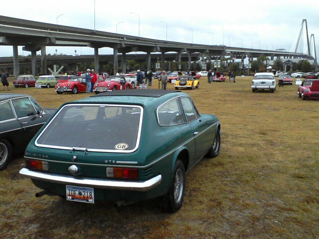 Car Show vs Hurricane Sandy - Page 1 - USA & Canada - PistonHeads - The image captures a vintage car rally held in a lush, verdant park. The focal point is a green British MGB sports car parked on a grassy area under an overcast sky. The MGB is surrounded by a variety of other classic and vintage cars, all sizes and colors, laid out in neat rows. A crowd of people can be seen milling about, admiring the vehicles. In the background, a large suspension bridge arches over the park, adding an interesting architectural element to the scene.