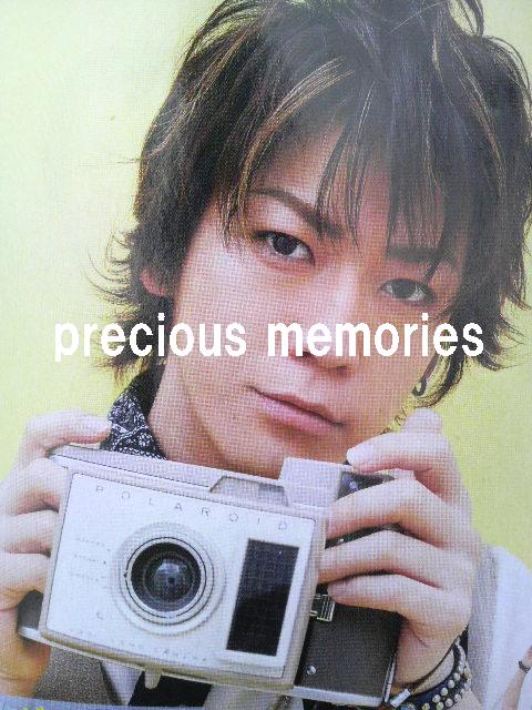 The image displays a person with expressive eyes and spiky hair. They appear to be holding a camera in their hands with one hand, while the other hand is near their shoulder or face. The person is wearing a light-colored shirt and possibly a patterned scarf. A charming detail is that a small smile is present, suggesting a pleasant demeanor. The background is out of focus, allowing the subject to be the main focus of the photograph.