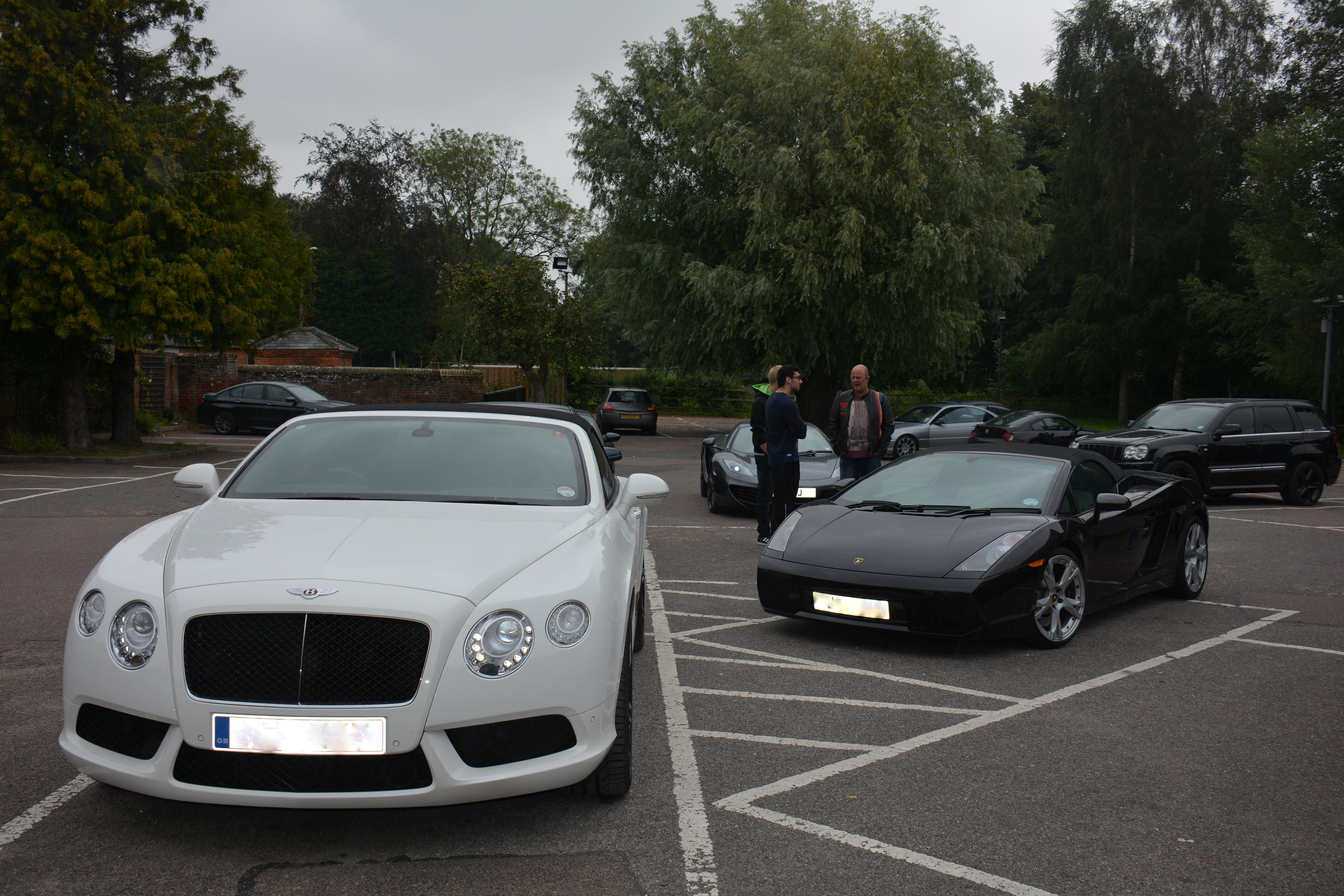 Clophill supercar meet 17th Aug - Page 2 - Herts, Beds, Bucks & Cambs - PistonHeads - The image depicts a parking lot filled with various luxury and convertible vehicles, including a prominent Bentley. There are several people standing around the cars, engaged in conversation and admiring the cars on display. The setting suggests a luxurious event, and the overcast sky provides a neutral backdrop for the subjects in the foreground. The scene is outdoors with trees and foliage in the background, indicating a park or a similar location adjacent to the parking lot.