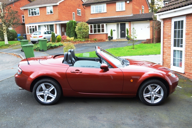 Will I ever find a MK3 in this specification? - Page 1 - Mazda MX5/Eunos/Miata - PistonHeads - The image shows a vibrant red sports car parked on the driveway of a residential house. The car, a convertible, is a two-door model with a visible top closure that suggests it's a soft top convertible. The side view and rear view mirrors indicate it's fully equipped with necessary safety features. The house in the background is a two-story building with a brick facade, featuring a double garage and a front yard with some greenery. The image has a slightly blurred effect, particularly around the edges, which gives it a tranquil, soft-focus quality, contrasting with the vivid red of the car.