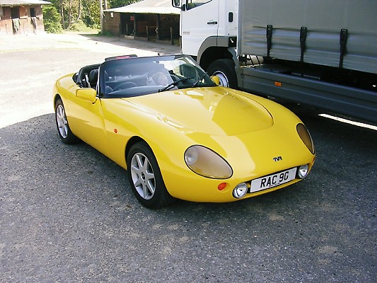 Lets see a picture of your classic(s) - Page 10 - Classic Cars and Yesterday's Heroes - PistonHeads - The image depicts a bright yellow Tran sports car, which is a targa top type of vehicle, positioned next to a large white delivery truck. The car is depicted from a front three-quarter perspective, allowing a clear view of its design, which features two doors and two seats. The word "TVR" on the grill of the car is visible, indicating its brand. The car is parked on a paved area which appears to be a road or parking lot with a gravel surface.