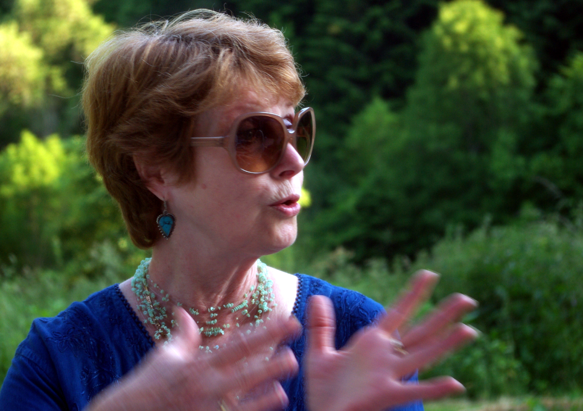 In the image, a woman dressed in a blue shirt and sunglasses is standing outdoors. Her hands are stretched out as she passionately talks, stunning the viewer with her gestures. She is surrounded by a serene natural setting, which includes trees and bushes. The colors in the image are vibrant, including the blue of the woman's shirt and the mix of greens and browns from the foliage and ground. The setting appears peaceful and tranquil, providing a calm backdrop to the woman's dynamic expression.