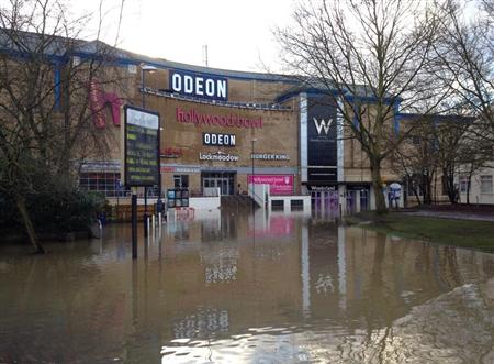 A large building with a clock on the side of it - Pistonheads - This image depicts a movie theater submerged in floodwaters. The evidence of the flood is largely covered by the water itself, but some landmarks remain visible, such as a large sign for the theater. The water's surface affects the view of the street, which runs alongside the theater. The surrounding area consists of trees and a clear blue sky in the distance, contributing to the overall scene of a flooded area alongside a building.