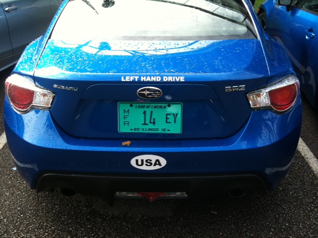 A car parked next to a parking meter - Pistonheads - The image shows a portion of a car parked in a parking lot. The car is painted a vibrant shade of blue and has a visible license plate. The license plate has a combination of letters and numbers with significant numeric values, and the text "Subaru" is also visible, indicating the make of the car. The environment suggests a rainy day given the wet appearance of the road surface and the splattered droplets visible in the background. The image captures the right side of the car, centered on the license plate.