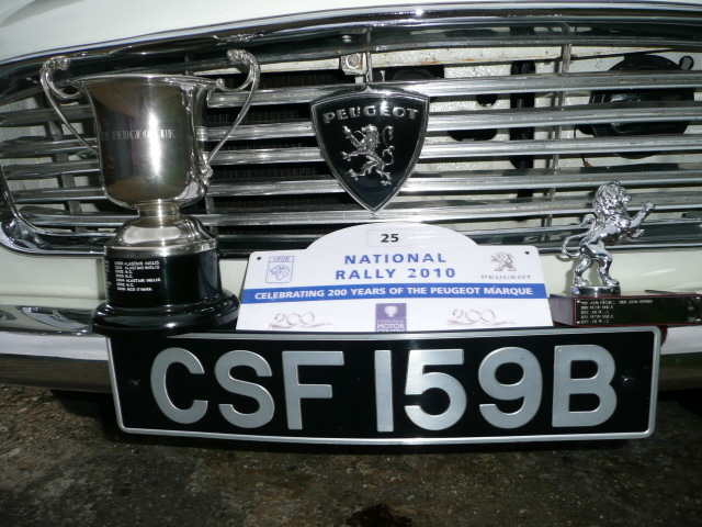 Correct font for 60's plates. - Page 1 - Classic Cars and Yesterday's Heroes - PistonHeads - The image features a vintage car with a notable crest on the grill. On the front of the vehicle, a racing trophy with the word "Bugatti" and the number "25" is mounted, signifying a significant achievement. Adjacent to the trophy, another trophy with the same "Bugatti" label and the number "42968" rests. In front of the car, there's a prominent black and white license plate with the acronym "CSF" and some additional numbers. A sign is also visible in front of the vehicle, commemorating the "National Rally 2010" and the "Pegoud Marque." The scene gives off a sense of automotive excellence and competition.