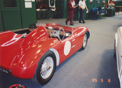 Early TVR Pictures - Page 35 - Classics - PistonHeads - The image features a classic red sports car with a white stripe, which appears to be a two-seater model. There is a man in a red suit standing next to the car, and another man in a white shirt is also visible in the background. The car is on a gray carpeted floor, and in the background, there's a white car parked. The visible part of the car displays accents such as chrome and polished metal, enhancing its sleek design.