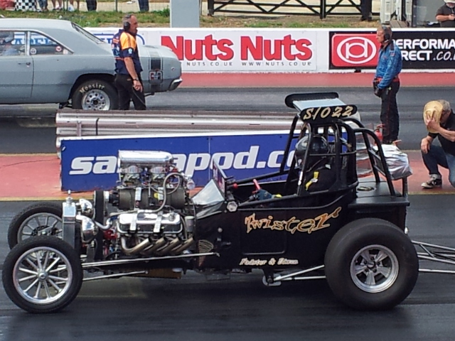 A man riding a motorcycle down a street - Pistonheads - The image features a powerful drag racing machine with a sleek black body, an engine with several visible components, large wheels, and gold accents. A race car is visible in the background with people standing around, one of whom is wearing headphones. The scene appears to be at a race track, with the racing machine ready at the starting line. The setting has a professional attitude, suggesting a competitive racing event.