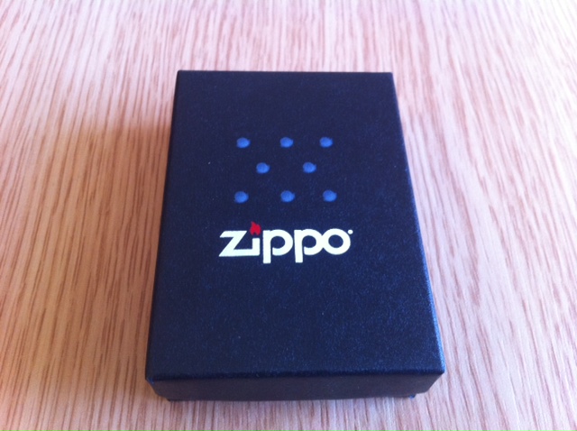 Show us your Zippo... - Page 7 - The Lounge - PistonHeads - The image showcases a product, specifically a box, placed on a wooden surface. The box is rectangular and predominantly black in color. An interesting feature is its textured top, which has a blue four-by-four grid pattern. A white oval logo of "ZIPPO" is displayed in the center of the lid. The overall presentation of the product is straightforward and does not contain any additional objects in the background. The lighting appears to be even, casting minimal shadows, highlighting the features of the packaging.