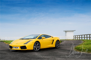 Pistonheads Locations Photoshoot Lambo - The image features a yellow sports car positioned on a road curving into the distance. The car is sleek and shiny, possibly a Ferrari, as indicated by the red and black detailing on its side. The backdrop consists of a blue sky and a vast expanse of green grass, with a white structure visible in the background. The overall mood of the image is dynamic and vibrant, suggesting speed and freedom.