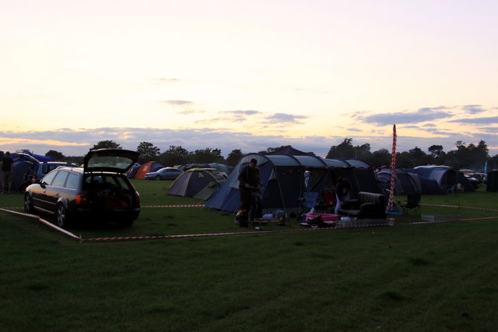 Previous Experience of Camping at Goodwood - Page 1 - Goodwood Events - PistonHeads - This image depicts a serene nighttime scene in a field that serves as a campsite. There are several tents spread across the grass, some occupied and others empty. A few cars, including two with their trunks open, are parked on the edge of the campsite, suggesting that campers may be in the process of settling in or have just arrived.

The field is quite open with a few bare trees dotting the landscape. A person can be seen in the distance, near one of the tents, possibly attending to tasks related to camping.

Above the field, the sky is dark and devoid of stars, indicating that it is likely nighttime. Despite the activity on the ground, the atmosphere remains overall calm and peaceful, reflecting the natural setting of the campsite.