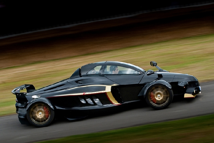 Watching Spain Pistonheads - This is a dynamic image of a black sports car with exterior highlights in gold and red. The car is positioned in the right lane of a street and appears to be in motion based on the blur effect captured in the photo. There are a couple of individuals inside the vehicle, likely enjoying a drive. The background includes a mix of elevated terrain with patches of grass and a horizon line that suggests the setting might be near the coast.