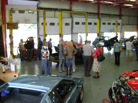 Pictures from Paul Roach's Open Day - Page 1 - Classic Cars and Yesterday's Heroes - PistonHeads - The image is a photograph taken inside a large, well-lit warehouse or garage with visible exposed flues or pipes on the ceiling. The floor is concrete, and the outfit of the people suggests a casual or social gathering, potentially a car show or convention. There are several cars on display, including a compact sports car with an open hood and a motorcycle, which appears to be of racing design with an exposed engine. People are standing around, mingling and observing the vehicles. There are also derby style cars parked on the far side of the photograph. The setting is industrial and characterized by robustness, with white walls and large garage doors that open into the partly cloudy daylight outside.