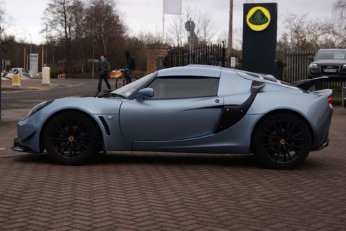 Exige intake winglets - Page 1 - Elise/Exige/Europa/340R - PistonHeads - The image depicts a futuristic, light blue over a darker blue, sports car parked on a brick surface, possibly a forecourt given the presence of a rounded sign with a logo that is partially visible and a person in the distance. It's a bright day, and the car appears to be a high-performance model, possibly a convertible given the presence of a retractable roof or a lighter roof panel. The car's modern design and color scheme suggest it is a contemporary or possibly conceptual vehicle.