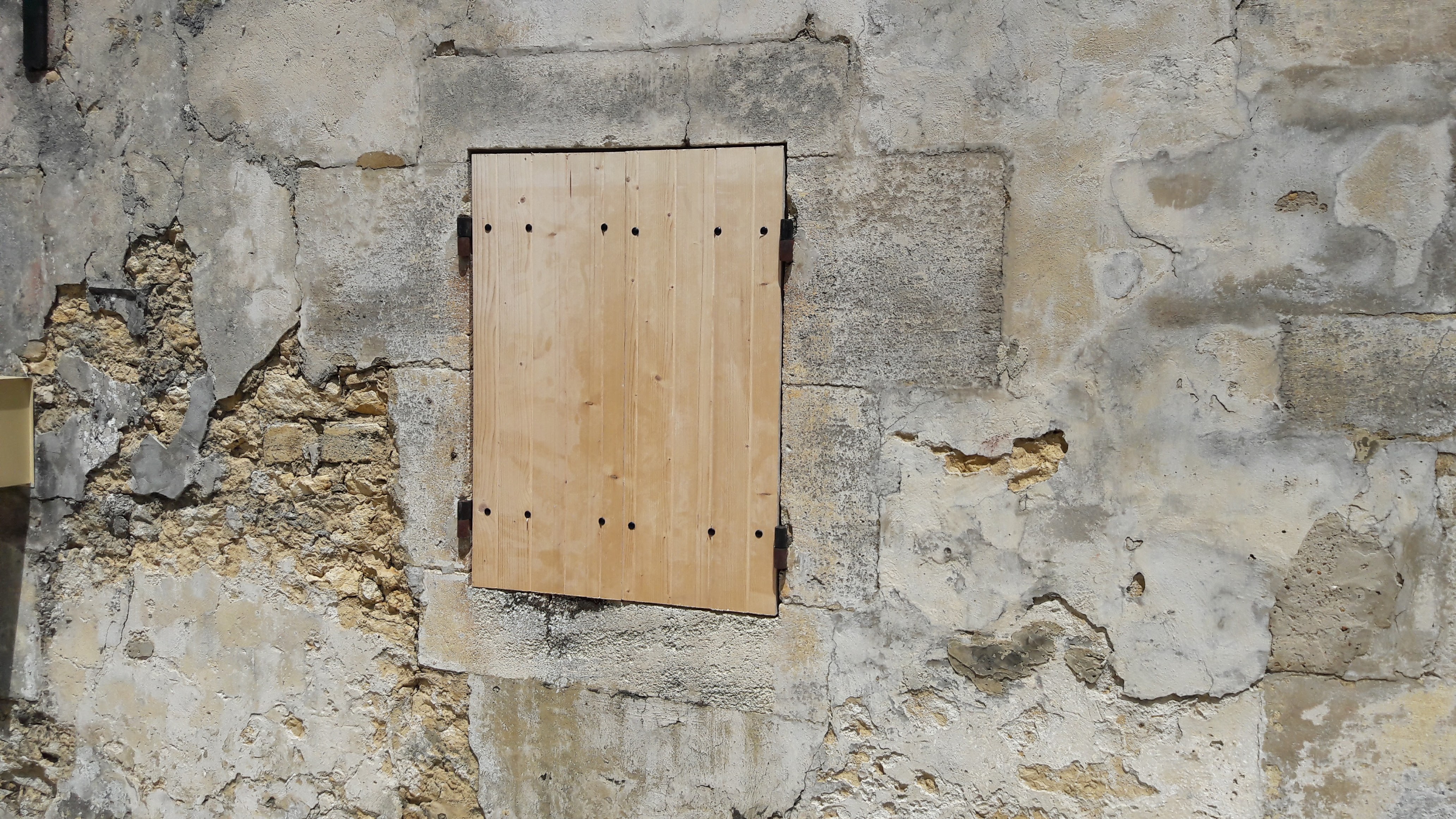 Our French farmhouse build thread. - Page 22 - Homes, Gardens and DIY - PistonHeads - The image shows a close-up view of a stone wall. Prominently featured is a wooden plank door, which appears to be mounted to the center of the structure. The door itself has eight small holes, indicating a possible locking mechanism or decorative embellishment. The surrounding wall shows signs of age or neglect, with flaking paint and sections of exposed brickwork, giving the building an older and dilapidated look.