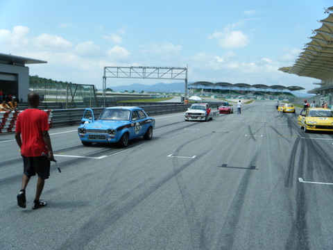 Afos Pistonheads Sepang - The image shows a racing circuit with several cars in a lineup, suggesting a racing event or a staged racing scene. In the foreground, there is a person in a red shirt walking towards the camera holding a pistol, which is not typical for a racing event and could be indicative of a movie scene or some creative purpose. The presence of the person with the gun adds a dramatic or action-oriented element to the otherwise standard racing scene.