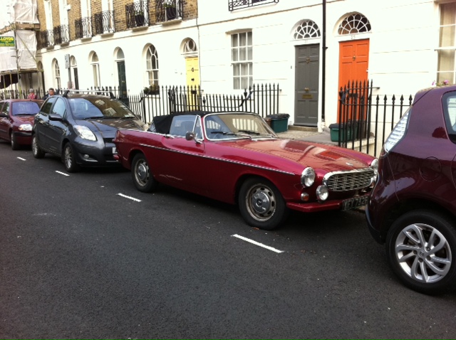 COOL CLASSIC CAR SPOTTERS POST!!! Vol 2 - Page 78 - Classic Cars and Yesterday's Heroes - PistonHeads - The image displays a scene of vintage cars parked along a street. The most prominent vehicle is a red sports car occupying two parking spots, with one spot behind another car and the second spot in front. The street is lined with buildings that look like residential or commercial establishments, as suggested by the presence of various doors.

In front of the red car, there's a purple car parked, while further in the background, there are other parked cars, contributing to a neat and orderly parking arrangement. The cars vary in sizes and colors, adding to the diversity of the scene. There's a sidewalk visible next to the parked cars, indicating that this is a public street with pedestrian access. The overall atmosphere of the image is calm and orderly, with no apparent movement or activity occurring. The image seems to capture a moment of stillness in an urban environment.