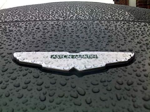 Your DB7 pictures. - Page 1 - Aston Martin - PistonHeads - The image showcases the wet surface of a car, which seems to be dripping with water. The car is a model of Aston Martin, as indicated by the logo placed in the center of the hood. The logo itself is adorned with water droplets, creating a shiny and reflective effect. The background is slightly blurred, indicating that the focus is on the front end of the car. The close-up perspective emphasizes the details and the shiny reflection of the car's surface.