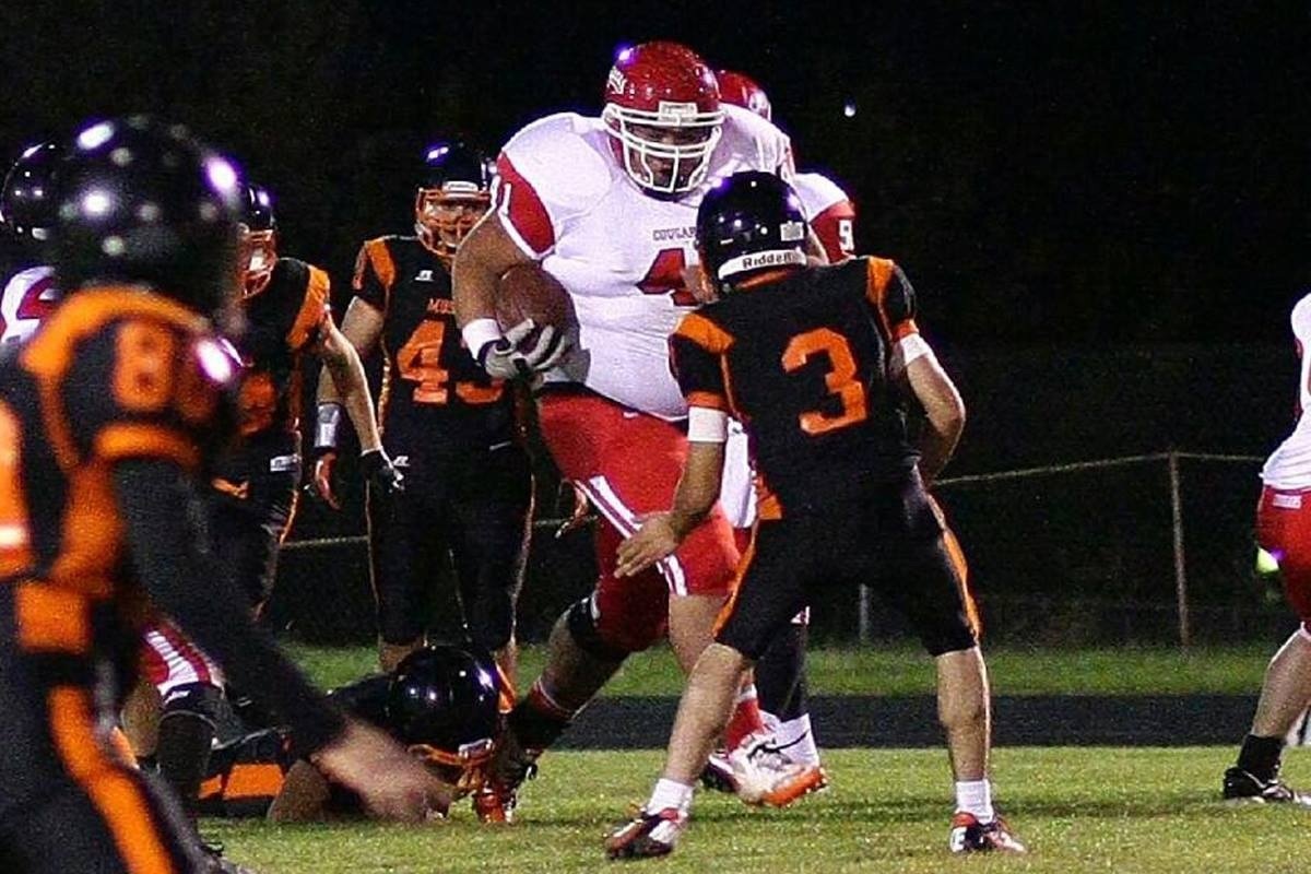 The camera can capture some fantastic moments IV - Page 88 - Photography & Video - PistonHeads - The image captures an intense moment in a high school football game. The action is concentrated around a large man in a white uniform, running with a football in his grasp, and smaller men in black uniforms trying to tackle him. The man in white has a determined look on his face as he holds onto the football. The black uniformed players are in various positions, some close and ready to make a play, while others are slightly further away. The image shows a clear delineation between the offensive team, represented by the player in white, and the defensive team, represented by the black uniformed players.