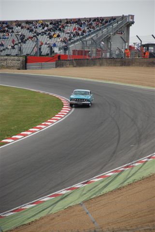 Hatch Brands Festival Masters Pistonheads - The image captures a dramatic moment on a race track. A blue car is racing around the curved track, making a sharp left turn. The car is in full motion, leaning into the curve with powerful intent. Numerous people can be seen in the background, likely spectators or other racers, adding to the sense of speed and competition. The grandstand in the distance is filled with onlookers, their presence underscoring the significance and excitement of the event.