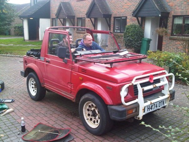 A red truck parked in front of a building - Pistonheads - The image captures an urban scene featuring a small, red SUV parked on a red brick sidewalk. The vehicle has a white roll bar visible on its roof. Inside the car, two individuals can be seen, both wearing jackets and seated on the back seat, seemingly engaged in a conversation. On the ground next to the car, there's a bottle of soda and a couple of random items. The background reveals a residential area with a building, a hanging flower planter, and a trash bin visible in the distance.