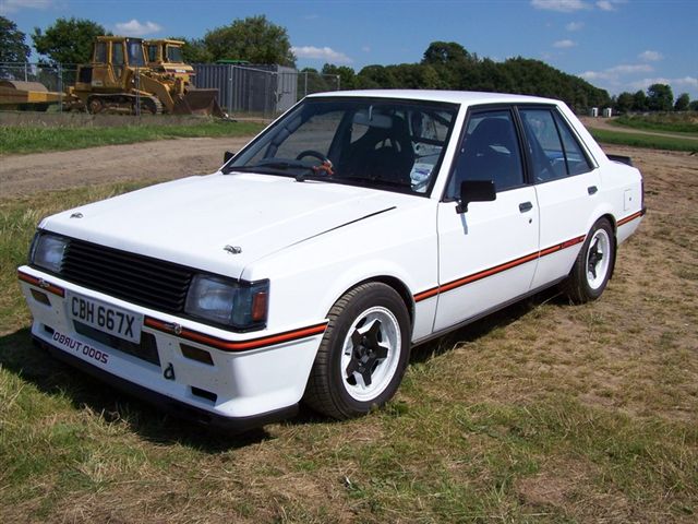 Turbo Pistonheads Lancer - The image features a striking white vintage car, identified by its license plate as a CT67X. The vehicle is parked on a grassy area, with a clear sky serving as the backdrop. A construction vehicle is seen in the background, suggesting that the area might be under development or that the car could be part of a car show. The contrast between the sleek vintage vehicle and the robust construction equipment adds an interesting dynamic to the image.