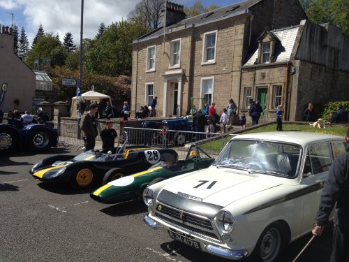 A group of people riding on the back of a truck - Pistonheads - The image depicts three vintage race cars parked side by side on a street. In the background, there is a small group of people standing and chatting. To the right, a man is holding the leash of a dog walking by. Further down the street, there's a white house with a green door and a cobblestone wall, along with another blue car. The scene gives off a nostalgic and relaxed ambiance, possibly indicating a small auto show or a gathering of car enthusiasts.