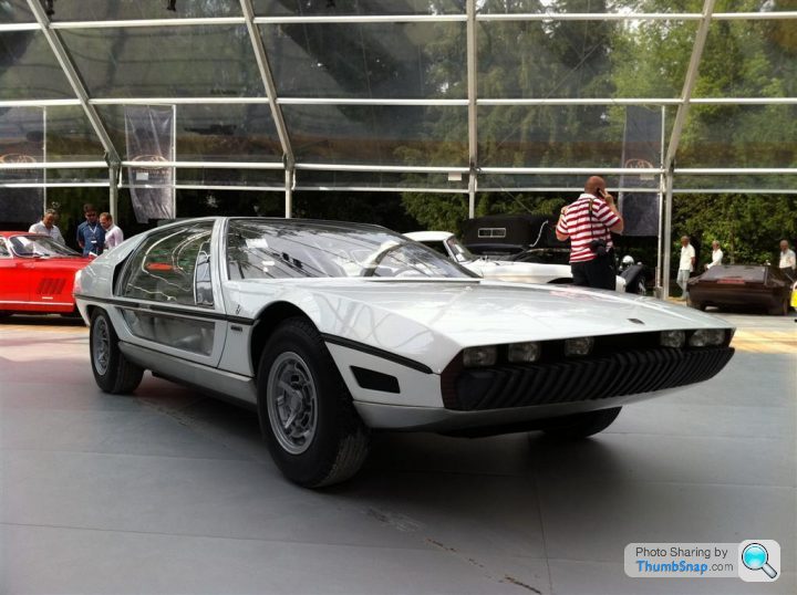 Pistonheads - The image showcases a man and a car in close proximity. The man, donned in a distinctive red and white striped shirt, appears to be engaged in a conversation on his cell phone. The silver car, which is a unique design that resembles a white Mazda, is parked under a glass pavilion, giving the car a sense of prominence. In the background, there are other people and cars, suggesting this could be a car show or a similar event. The man also has a baseball cap, possibly signifying his interest in sports or his role as an attendee or participant at this event.