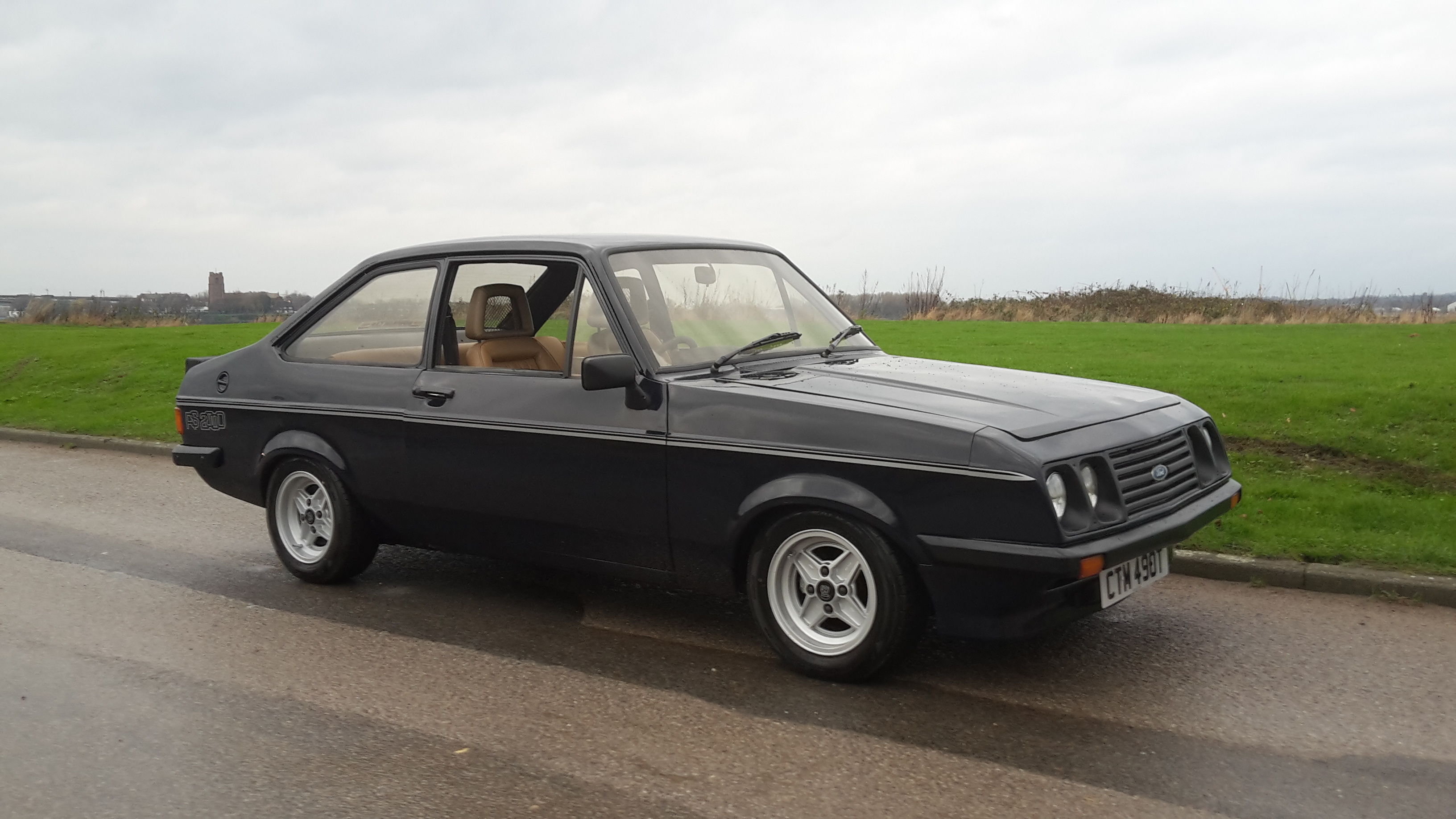 RS2000 MK2 RESORATION - FOR THE BRAVE / STUPID - Page 6 - Classic Cars and Yesterday's Heroes - PistonHeads - In the image, a black vintage car is the central focus. It's parked on the right-hand side of a wet road, which reflects light and adds a shimmering effect to the wet pavement. The car appears to be in good condition, and its classic design stands out against the backdrop of a relatively uneventful street. The road seems slightly curved, with no other visible vehicles or significant landmarks, giving the impression of an empty, poignant street. The overall scene is quiet and subdued, with the car as the sole point of interest.