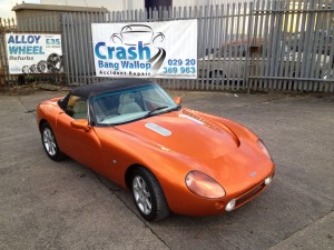 new colour - Page 1 - Griffith - PistonHeads - The image shows a vibrant orange sports car with its roof down, parked on a concrete surface next to a chain-link fence. A sign is placed in front of the car on the grass, suggesting that it might be on sale or for display. The sign has text that includes "Crash", "Bang", and "Wallop", possibly indicating the services or products associated with the car. In the background, there are other vehicles and a partially visible sign advertising "Alloy Wheels" and "Accident Repairs".