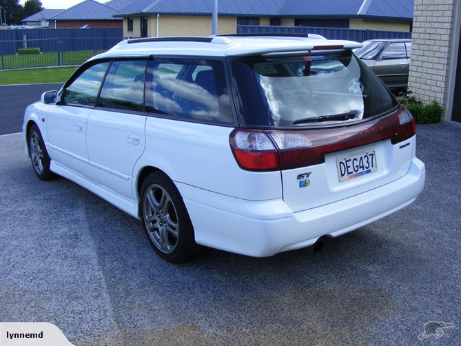 My new Legacy - Page 1 - Subaru - PistonHeads - The image displays a white automobile parked on a driveway. The car features a red tail light area, and there is a license plate visible at the back. The vehicle has a hatchback style, and it appears to have a full-sized spare tire mounted on the trunk lid. The surroundings show a house with a blue roof in the background; there is a fence, a lawn, and a paved surface. In the bottom right corner, there is a watermark with the signature "Lynne" and a graphical logo.