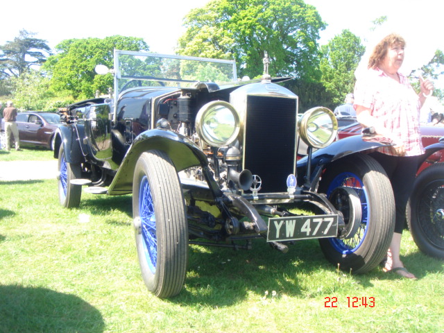 Old Warden car show - Page 1 - Classic Cars and Yesterday's Heroes - PistonHeads - The image depicts a vintage black car with thick white stripes and a prominent large radiator grill, characterized by round headlights and a bonnet with a ribbed texture. The car is positioned in a lush green field with trees in the background. On the left side of the photograph, a woman is visible, standing close to the antique car. The setting appears to be a casual outdoor event. The timestamp on the image reads "22 12:43". The car has the registration number YJW 477.