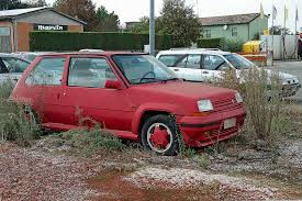 Classics left to die/rotting pics - Page 362 - Classic Cars and Yesterday's Heroes - PistonHeads - The image depicts a scene in a parking lot. The primary focus of the photograph is a small red car parked on a patch of grass and dirt surrounded by weeds. To the right and in the background, there are various vehicles, including other cars and a truck. These vehicles are parked on pavement, contrasting with the unpaved area where the red car is parked. The background also features a green building with a sign that includes the word "harris," all visible under an overcast sky. The style of the image is a straightforward, non-stylized photograph intended to depict the stated scene clearly and without embellishment.