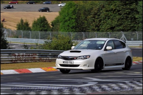 Pistonheads - This image captures an exciting moment on a winding race track. At the center of the frame is a sleek, white sports car with a black spoiler, emitting a sense of speed and power as it maneuvers the track. The car's license plate, clearly visible, reads "M33 TSI." The track itself is predominantly black, contrasting with the vibrant green of the surrounding trees and shrubs. A chain-link fence runs along the base of the track, and beyond it, buildings and cars can be seen in the distance, adding a touch of the real world to this racing scene. The overall scene is a thrilling blend of high-speed driving and scenic backdrop.