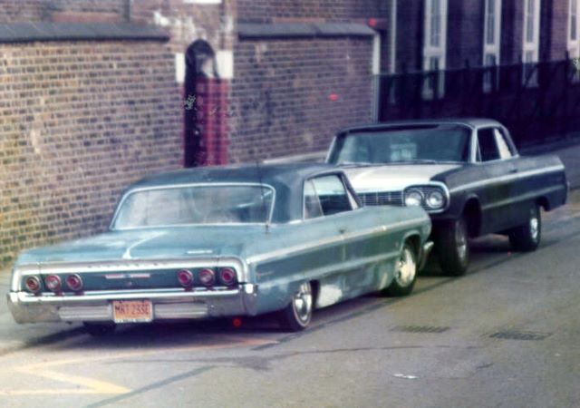 1964 Impala - Page 1 - Yank Motors - PistonHeads - In the nighttime scene, two vintage cars, one black and the other silver, are parked next to each other on the side of a road. The black car is parked behind the silver car, facing us, while the silver car is parked in front, facing away. Both cars exhibit classic styling reminiscent of the early 1960s, adding a touch of nostalgia to the scene. The presence of a brick wall and a contrasting refined wall in the background suggests an urban setting. A solitary figure in the background seems to be observing the scene, contributing to the quiet atmosphere of the image.