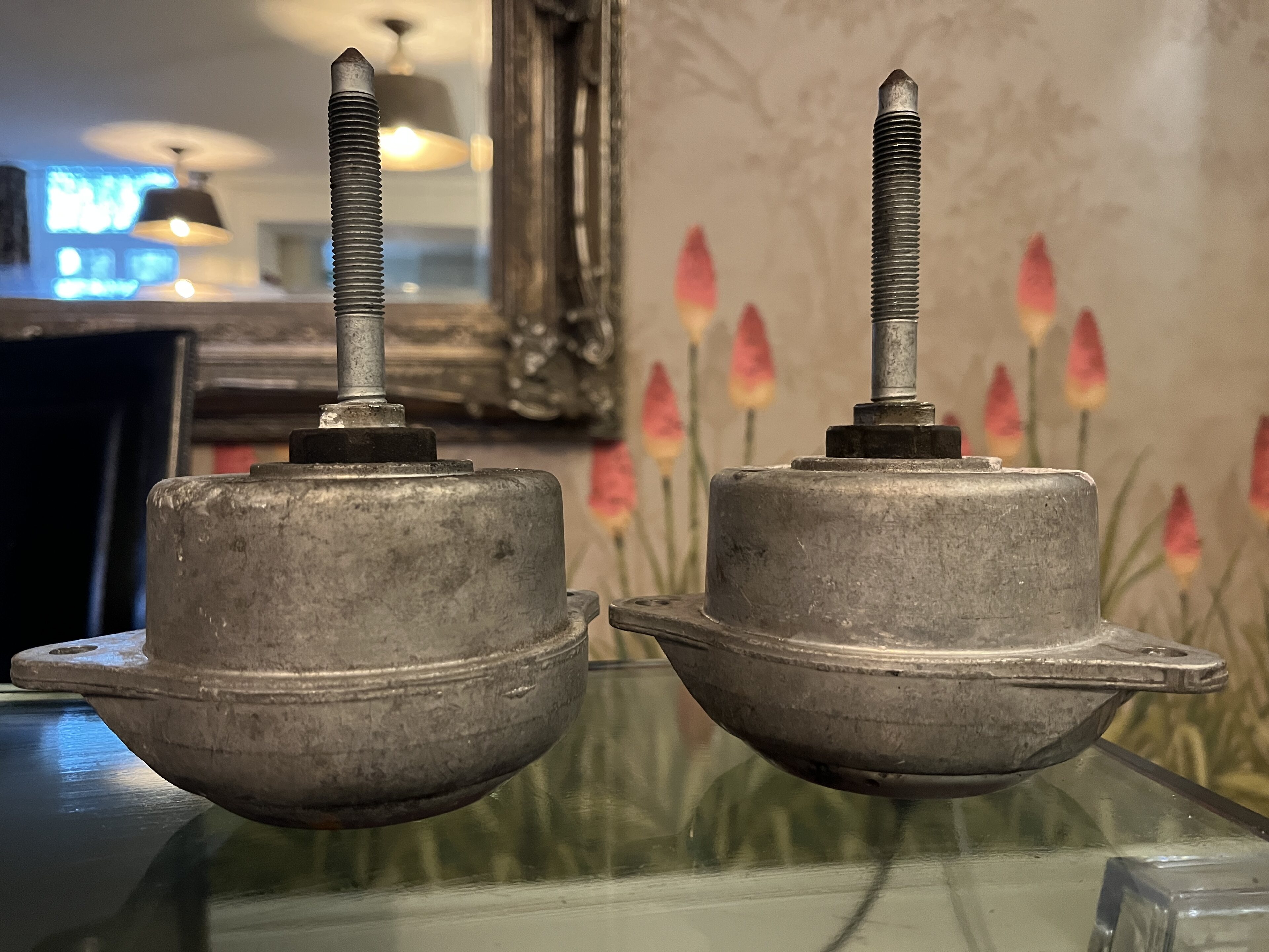 Pistonheads - The image shows two round, metal objects placed on a table. They appear to be a part of some machinery or device, given their structure and the presence of bolts and screws in them. The background includes a mirror and a framed picture with a floral motif on the wall, suggesting an interior setting, possibly a home or office. There is no text present in the image.