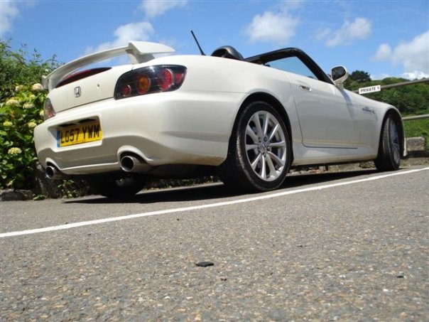 White S2000 - Page 1 - Honda - PistonHeads - The image shows a white, two-door convertible car parked on the side of a road. The car is faced towards the left side of the image, showcasing the wheels and the back half of its body. In the background, there are trees and a clear sky, suggesting the photo was taken during the day. The car appears to be a hardtop convertible model, possibly an Honda, as indicated by their logo on the trunk.