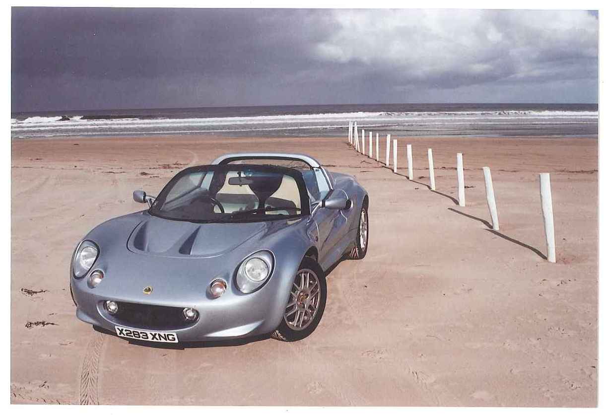 The big Elise/Exige picture thread - Page 16 - Elise/Exige/Europa/340R - PistonHeads - The image features a silver sports car parked on a sandy beach near the ocean, with a series of white poles, possibly for vehicles, positioned in the background. The car exhibits sleek lines and a curved windshield, indicating its sporty design. The beach stretches out towards the horizon, meeting the sky, suggesting the photo may have been taken at sunrise or sunset given the soft, diffused light casting a gradient on the sky. The ocean's water shows waves, indicating gentle surf conditions.