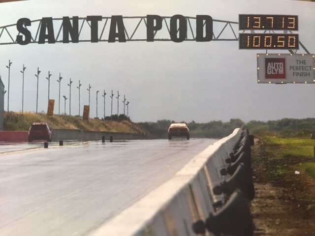 My Mach 1 showing a souped up Fiesta some dust - Page 1 - Mustangs - PistonHeads - The image shows a scene at Santa Pod race track, known for world championship drag racing. On the track, two cars can be seen with their exhaust pipes visibly raised, suggesting they are in the midst of a high-speed run. Above the track, there is a scoring board indicating times of 13.71 and 10.50 seconds for an event called "The AutoGlym Perfect Finish". In the distance, the race track gates read "Santa Pod Raceway" and there is a truck which could be part of the support crew or a barrier-enhancement vehicle. The sky appears overcast, and the lush greenery surrounding the track gives a sense of a well-maintained outdoor facility.