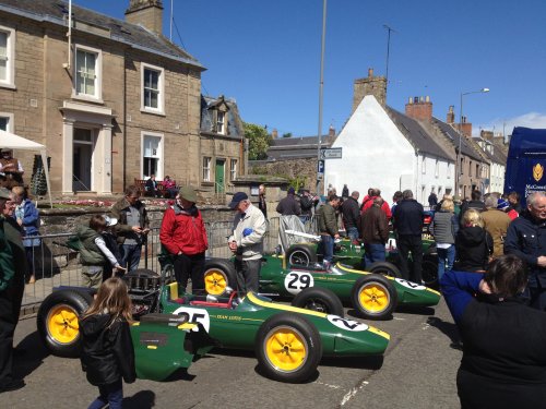 A large group of people on a city street - Pistonheads - The image showcases a vibrant scene on a sunny day. Two old-fashioned cars, painted in shades of green and yellow, are the main attraction. They are parked side by side on a sunlit street, with a crowd of people gathered around, admiring the vintage vehicles. The cars are adorned with large numbers 29, suggesting they might be vintage race cars or a part of a racing event. The crowd seems to be enjoying the spectacle in front of them, adding to the festive and lively atmosphere.